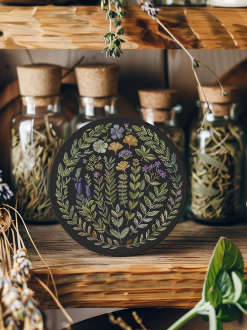 Circular ceramic coaster with black background featuring intricate botanical illustrations of various herbs, flowers and leaves in green, purple and gold colors with white ceramic edge and cork backing.